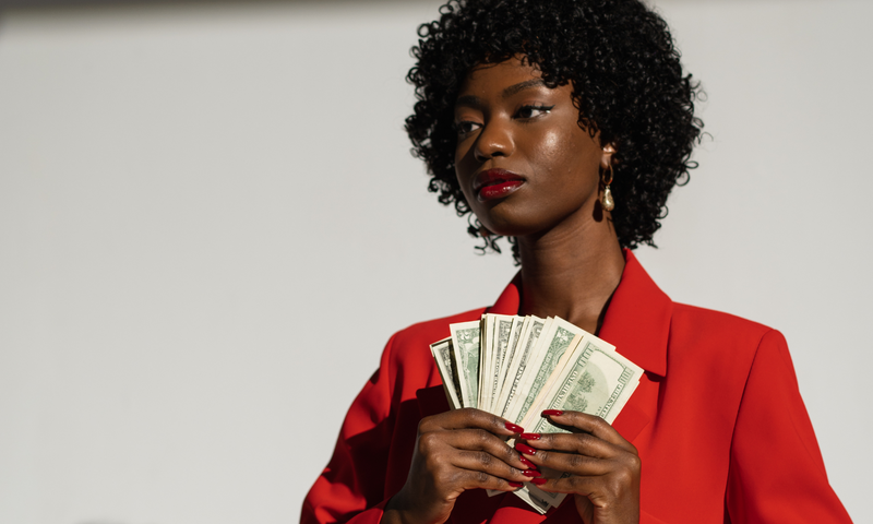 Confident black woman in red blazer holding dollar bills.