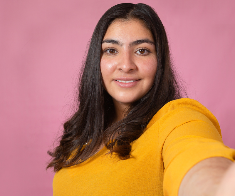 Latin woman taking a selfie, wearing a yellow shirt, with a pink background.