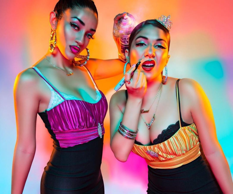 The Chonga Girls, in vibrant outfits and tiaras, pose playfully with makeup and a hairspray can in colorful lighting.