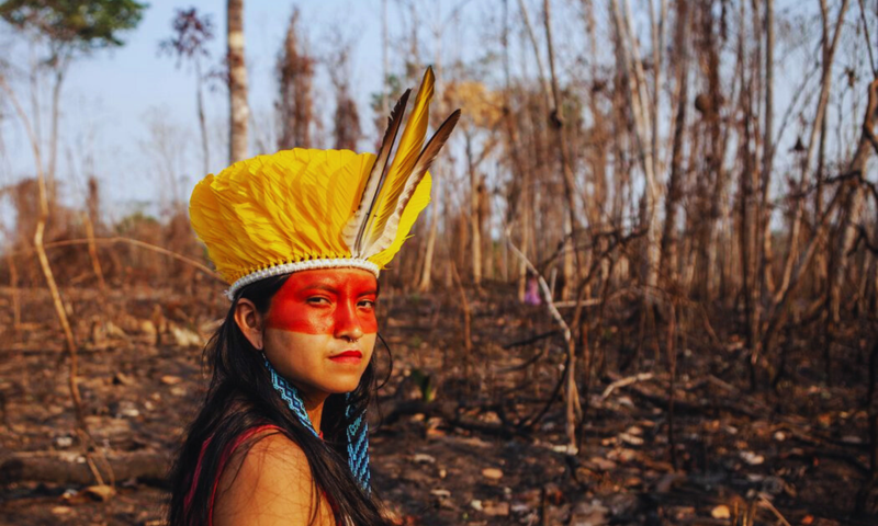 Indigenous woman from the Huni Kuin tribe with traditional face paint and feather headdress in a deforested area.