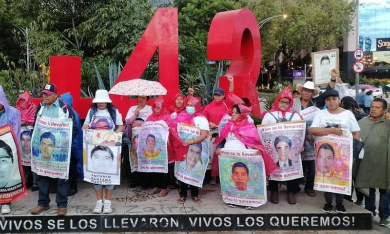 People protesting with signs by a red '43' sculpture, marking the Ayotzinapa students' disappearance.